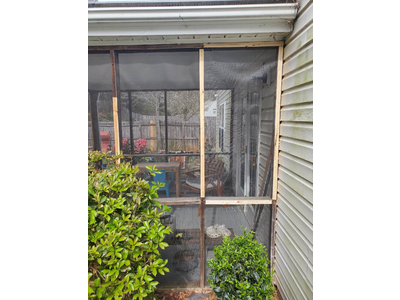 A screened porch with crude wooden framing. Some panels are chicken wire. The siding of the house is dirty.
